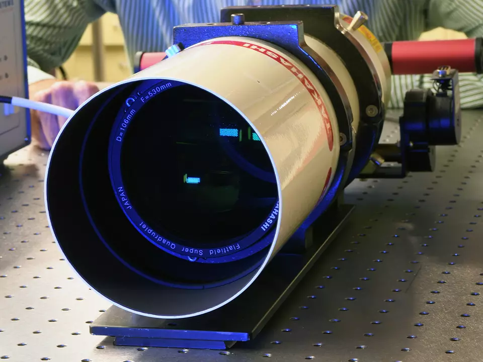 The dark opening of a small telescope is pointed toward the viewer as the instrument sits on a lab table.