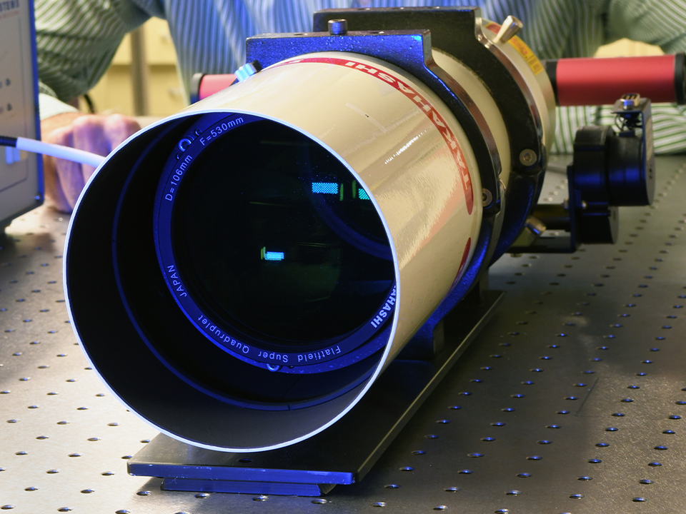 The dark opening of a small telescope is pointed toward the viewer as the instrument sits on a lab table.