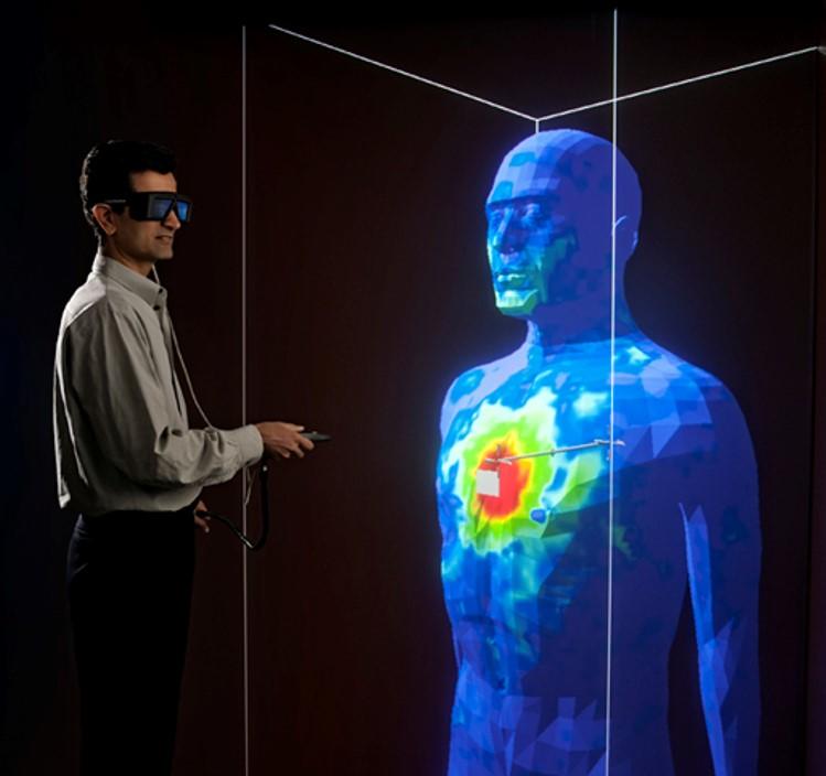 A researcher in safety glasses stands holding a remote control near a large blue model of a human head and torso with red marks on the chest. 