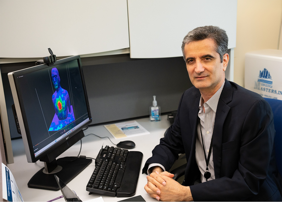 Kamran Sayrafian poses sitting at his office desk, with a computer monitor showing a colorized scan of a human head and torso. 