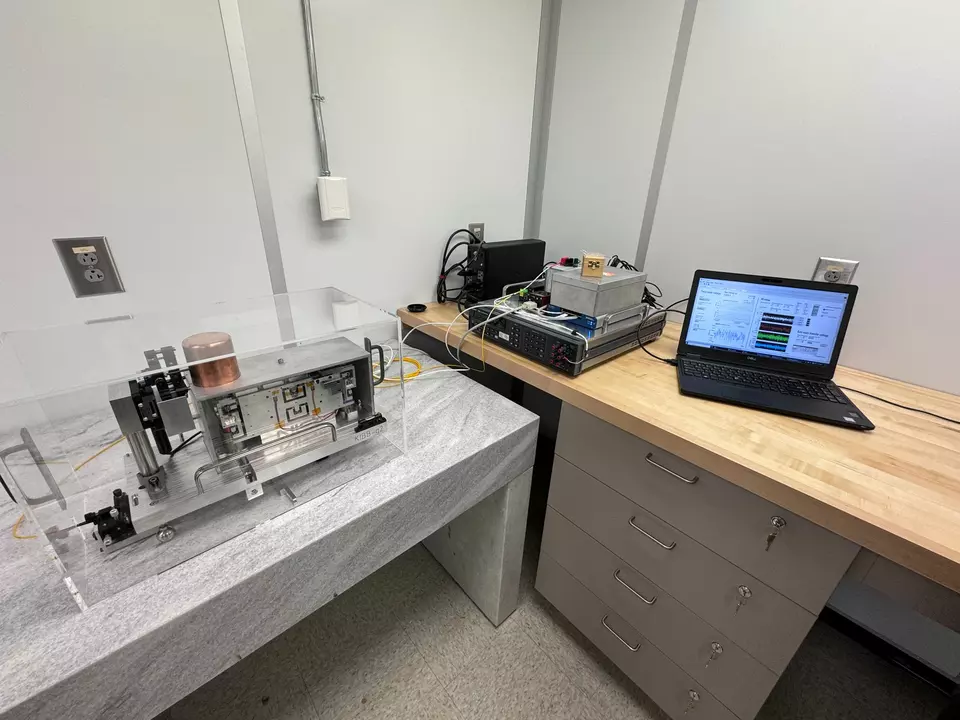 A scientific instrument sits inside a clear plastic box on a table near a desk with a laptop computer. 