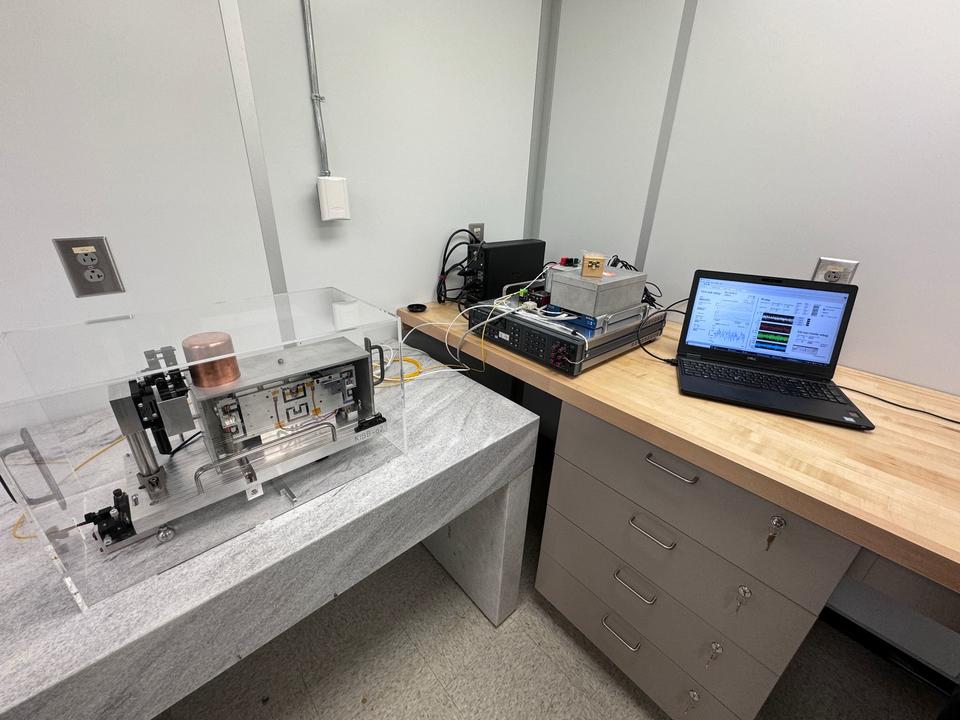 A scientific instrument sits inside a clear plastic box on a table near a desk with a laptop computer. 