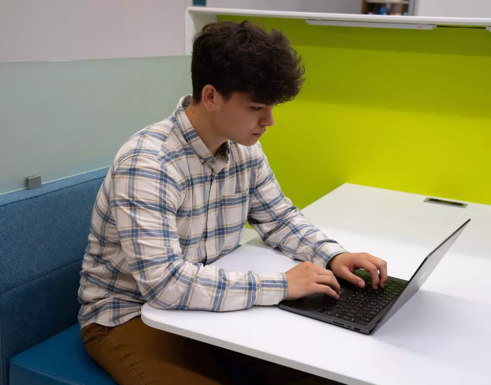 A man works on a laptop sitting at a white table in front of a brightly colored wall. 