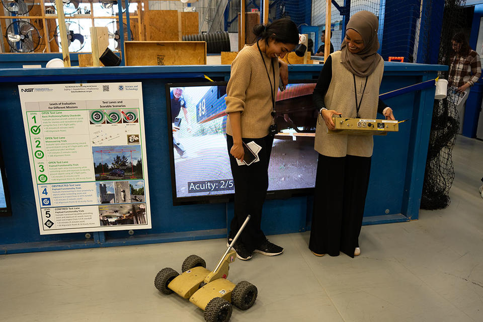 Two women stand in a lab space looking down at a wheeled robot. The woman on the right holds a large control panel.  
