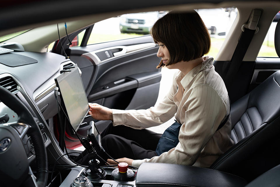 A woman sits in the passenger side of a parked car, looking at a large monitor attached to the dashboard.