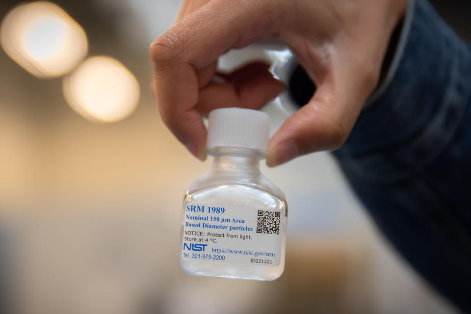 A hand holds a small, clear bottle full of a liquid solution with a label that says SRM 1989.