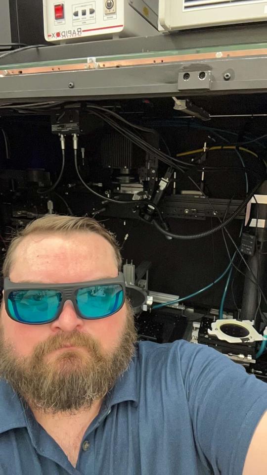 A bearded man wearing reflective safety glasses stands in front of an open cabinet full of scientific equipment. 