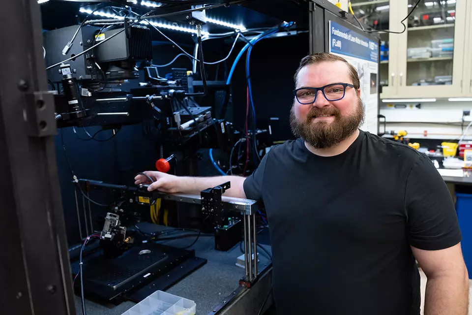 A bearded man poses smiling next to an open cabinet of equipment in a lab. 
