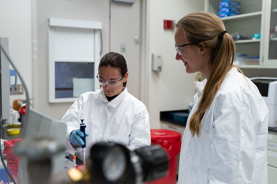A researcher in a lab coat and safety glasses uses lab equipment as another researcher looks on. 