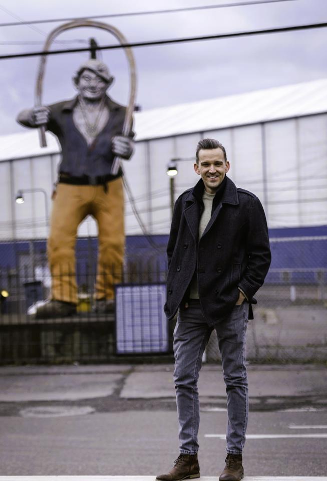 Andrew Iams poses smiling in front of a large statue of an ironworker outside a factory building. 