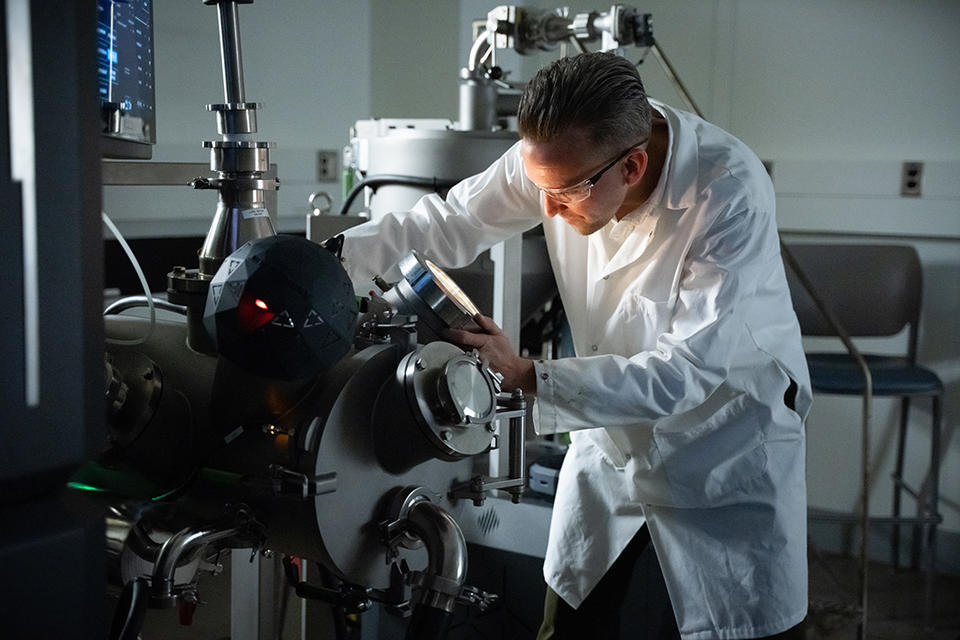 Andrew Iams wears a lab coat and safety glasses as he looks into a circular opening on a large piece of lab equipment. 