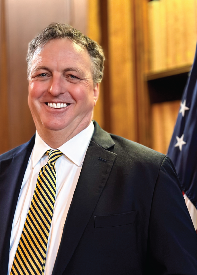 Photo of Mark Schmit standing in front of an American flag wearing a suit.