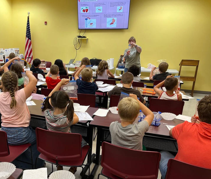 Sierra Miller stands in front of a classroom holding a ziploc bag. Kids are sitting at desks holding up their own bags.