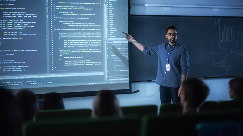 A young male teacher stands at the front of a darkened classroom, pointing at computer code on a large screen. 