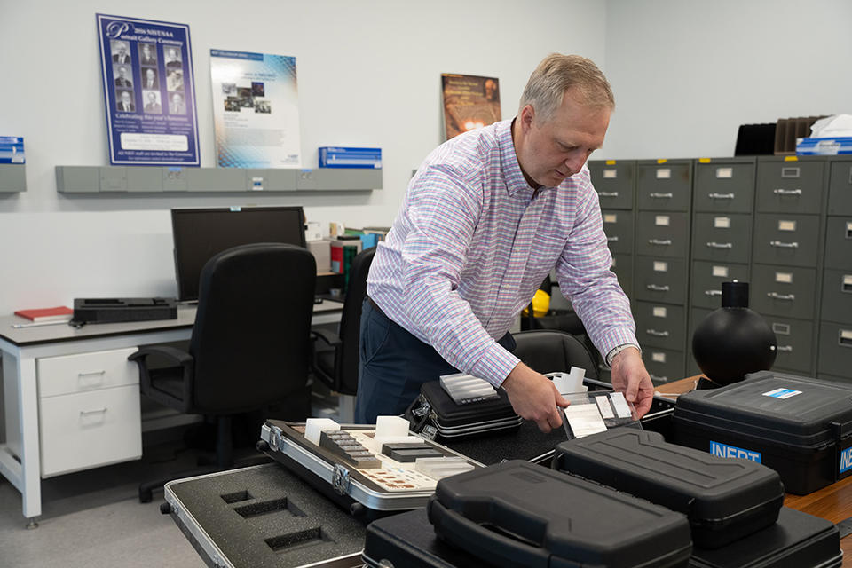In an office with a desk and filing cabinets, Jack Glover leans over a table holding black cases with customized foam inserts for equipment. 