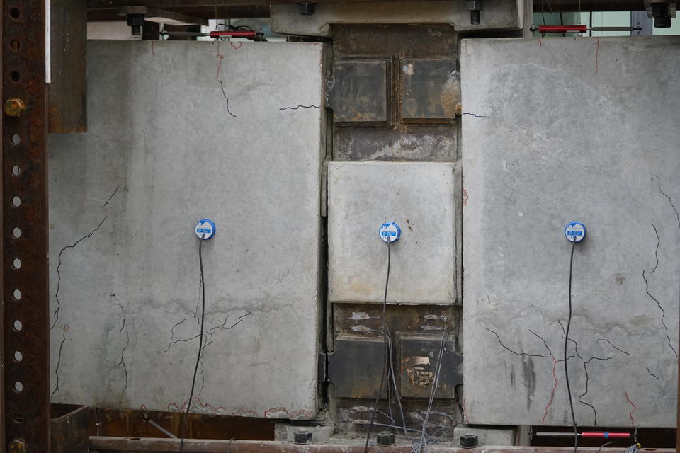 A wide view a concrete beam assembly inside a large metal frame. There are multiple small cracks in the concrete highlighted with Sharpie. One of the steel plates connecting the concrete pieces together is broken.