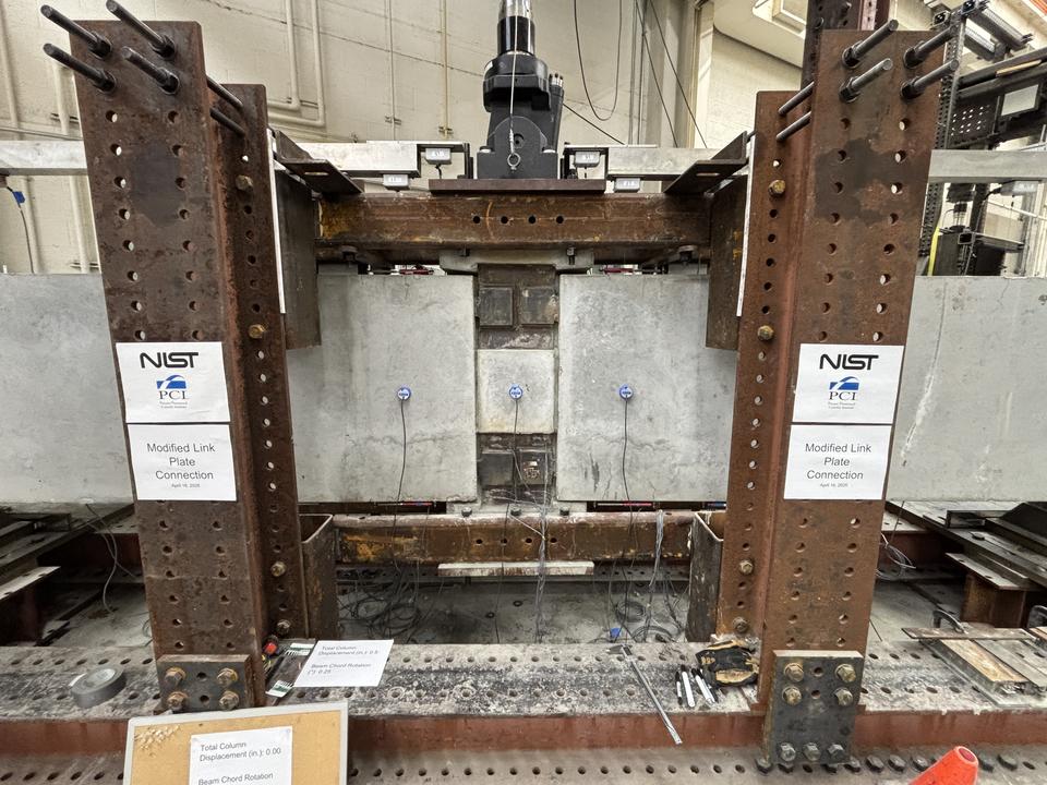 A wide view of a concrete beam assembly inside a large metal frame. A hydraulic arm is part of the metal frame, and it's posed to press down at a section in the middle where the concrete pieces connect.