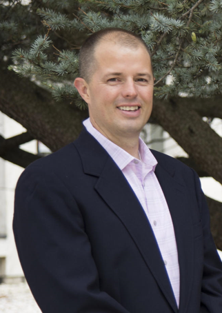 A man with short hair, wearing a blazer and collared shirt, standing outside, smiling.