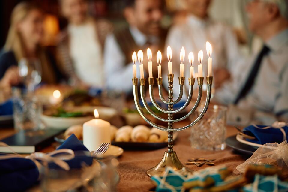 A lit menorah stands on a table surrounded by food and people in the background. 
