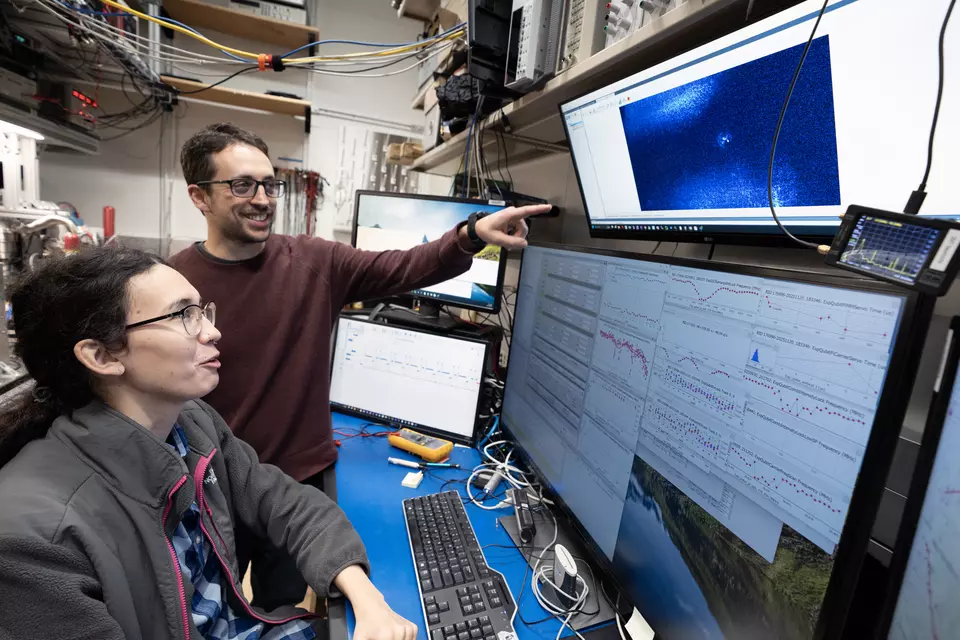 A woman with glasses and a long ponytail is seated at a computer with four display screens. A man with glasses stands beside her and points at the uppermost display screen. In the middle of that screen is a small blue glowing dot. The lower screens display data from the experiment.