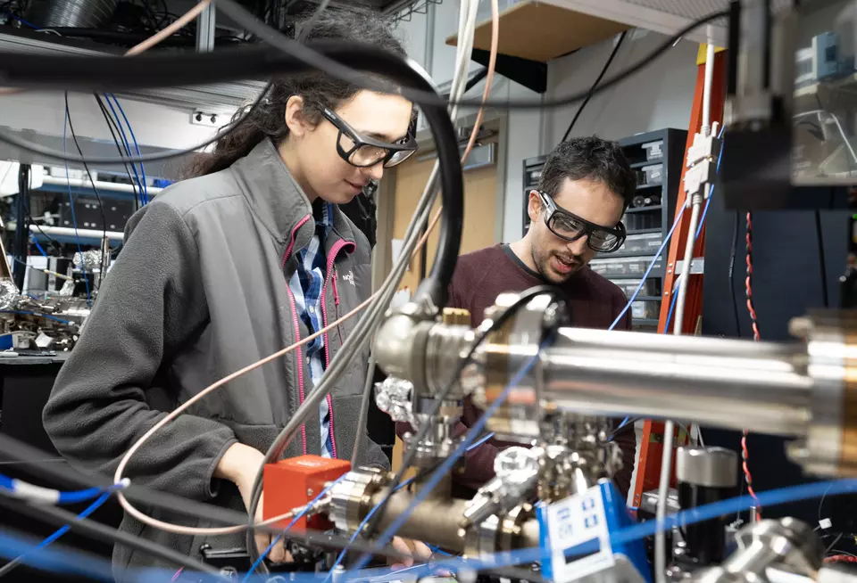 Two researchers wearing safety glasses are seen standing in the lab surrounded by wiring and electronics.