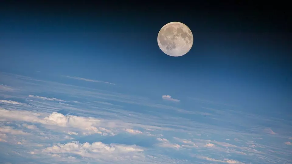 A full moon appears in the darkened upper part of an otherwise blue sky, with clouds below.. 