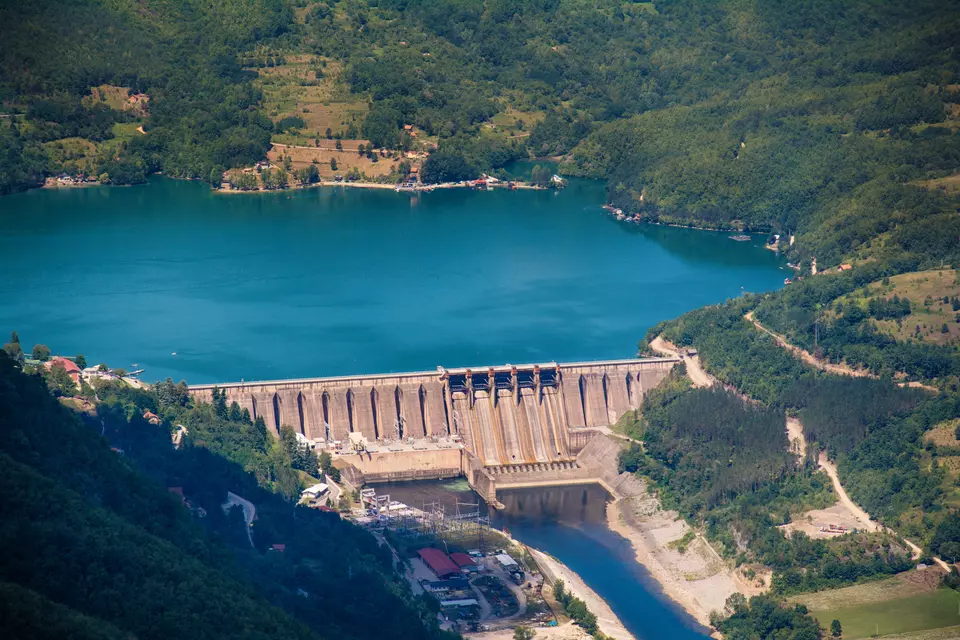 Shown from above, a large concrete dam holds back a lake of blue water surrounded by green hills. 
