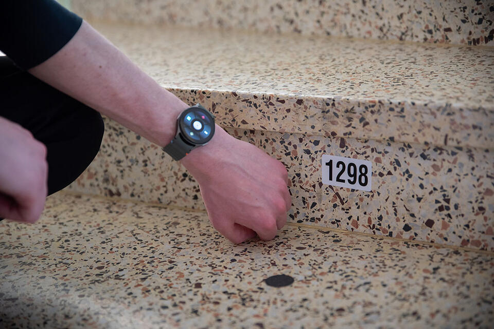A person wearing a specialized smartwatch gestures to a black dot sticker with a nearby numbered sticker on a set of stairs.
