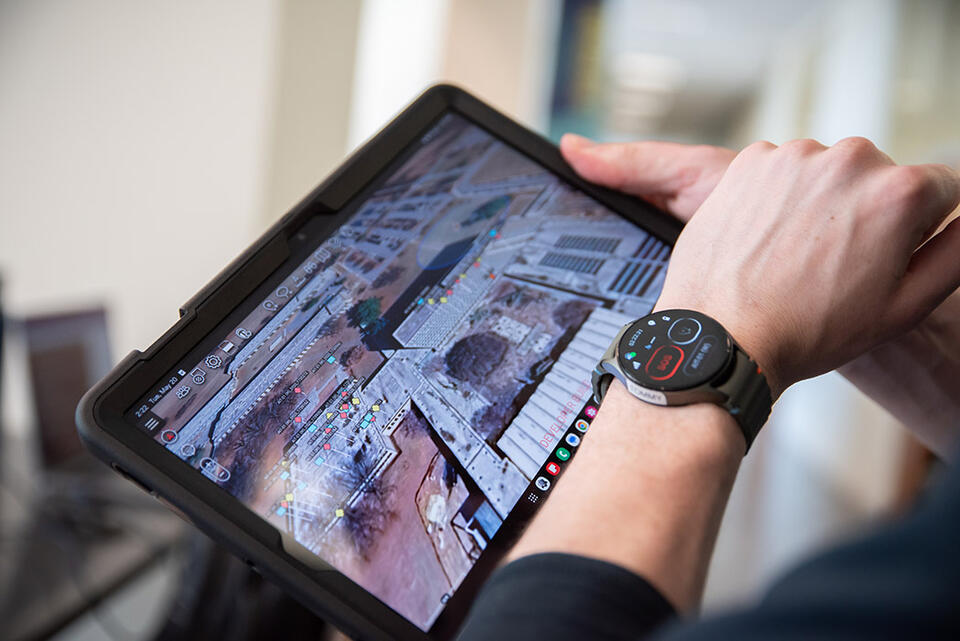 A person wearing a specialized smartwatch holds a tablet showing a photo of the NIST campus with colored dots marking certain locations. 