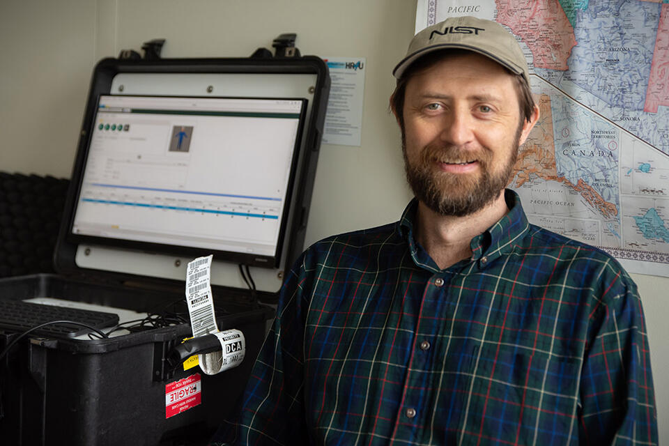 Gary Howarth poses for a photo in his office, wearing a NIST baseball cap, with a ruggedized laptop on the counter next to him and a map on the wall behind him.. 