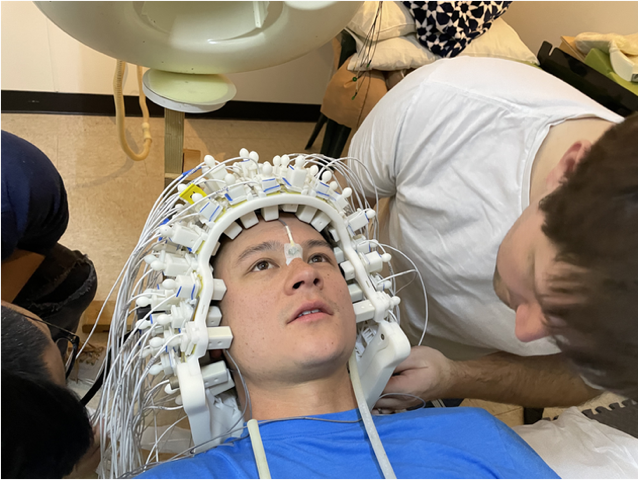 A researcher leans over a person wearing a white helmet made of many small devices and wiring.