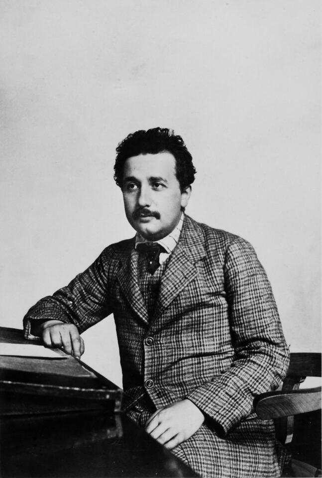 A young, dark-haired Albert Einstein poses sitting at a desk. 