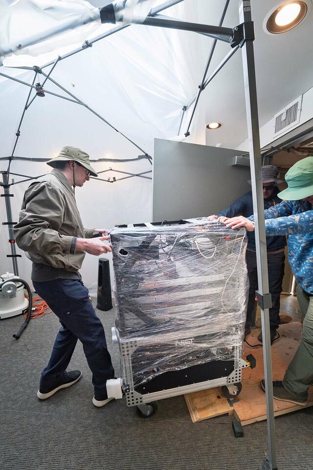 Researchers in wide-brimmed hats guide a shrink-wrapped cart of scientific equipment through a doorway. 