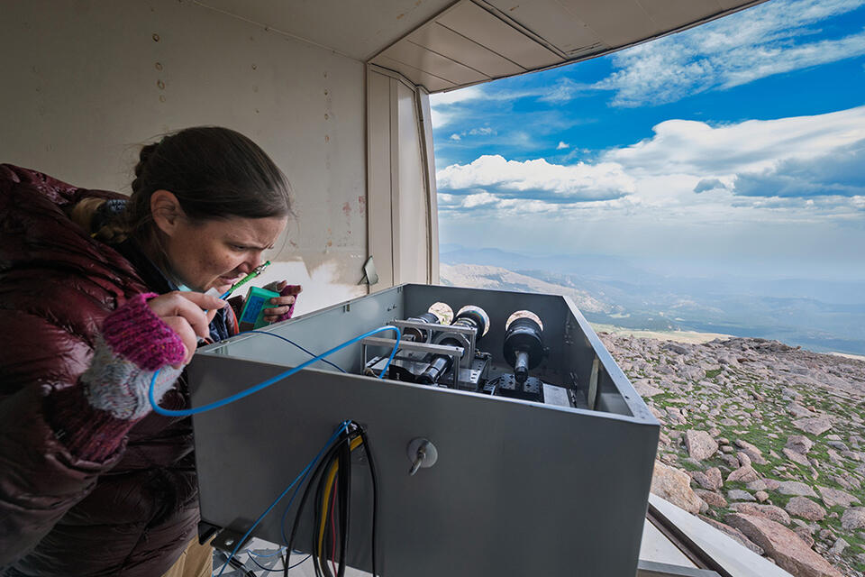 A researcher peers into a box of scientific equipment near an opening in the wall showing the view down the mountain. 
