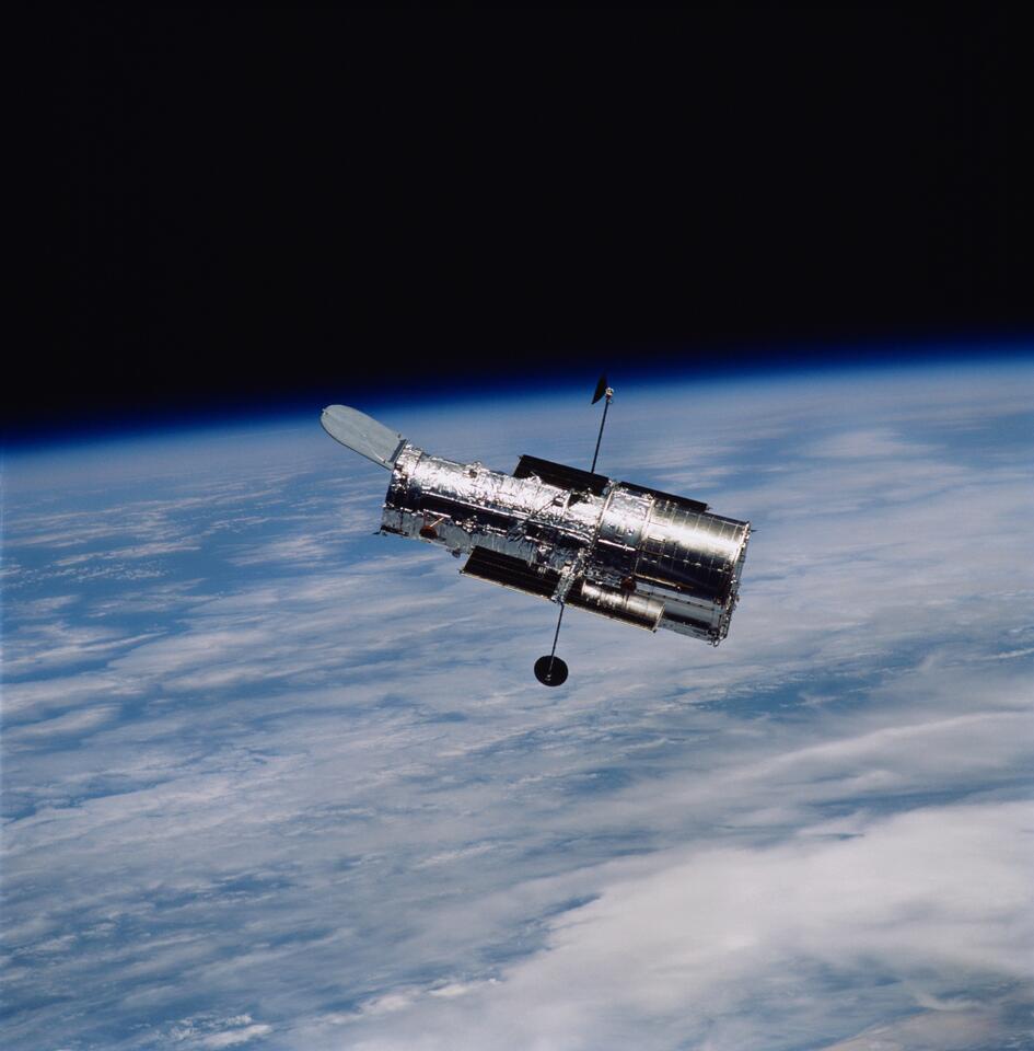 The Hubble Space Telescope (HST) is a floating silver cylinder with the backdrop of the horizon of Earth and the blackness of space.