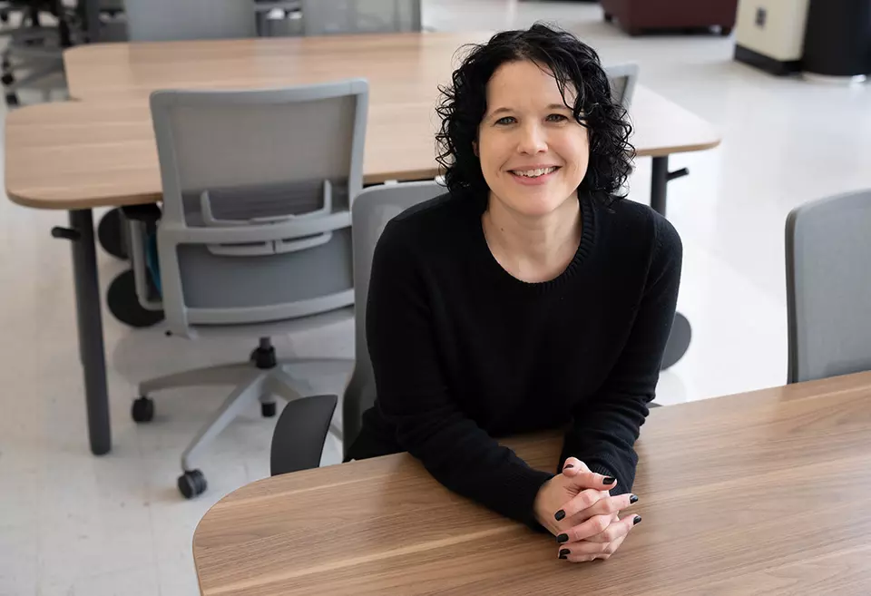 Kelly Sauerwein poses sitting at a table in the NIST library, leaning forward and smiling. 