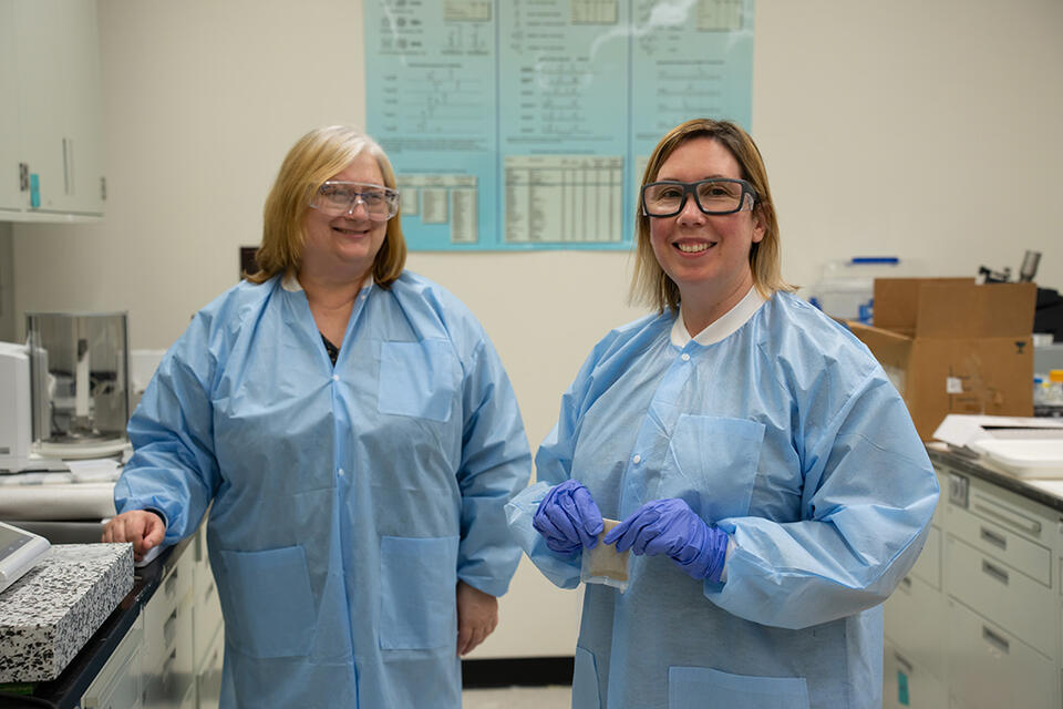 Two women in blue paper lab coats and safety glasses stand smiling in the lab. 