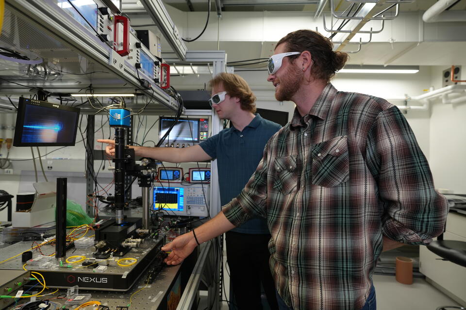 Two male researchers in casual clothes and protective eyewear stand in the lab, pointing at a screen hooked up to a laser science instrument. 