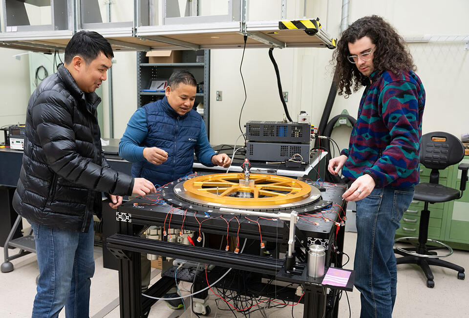 Three warmly dressed researchers stand around a scientific device with a large circular piece lying flat on top. 