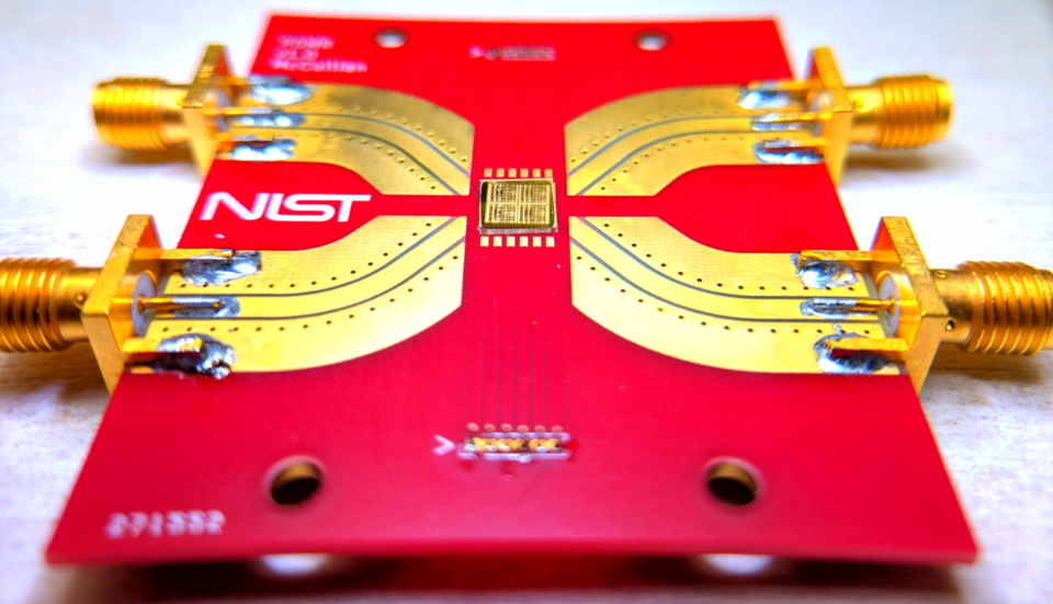A bright-red rectangle with a NIST logo holds a gold-colored chip in the center with metal paths leading to connectors at the edge of the rectangle. 
