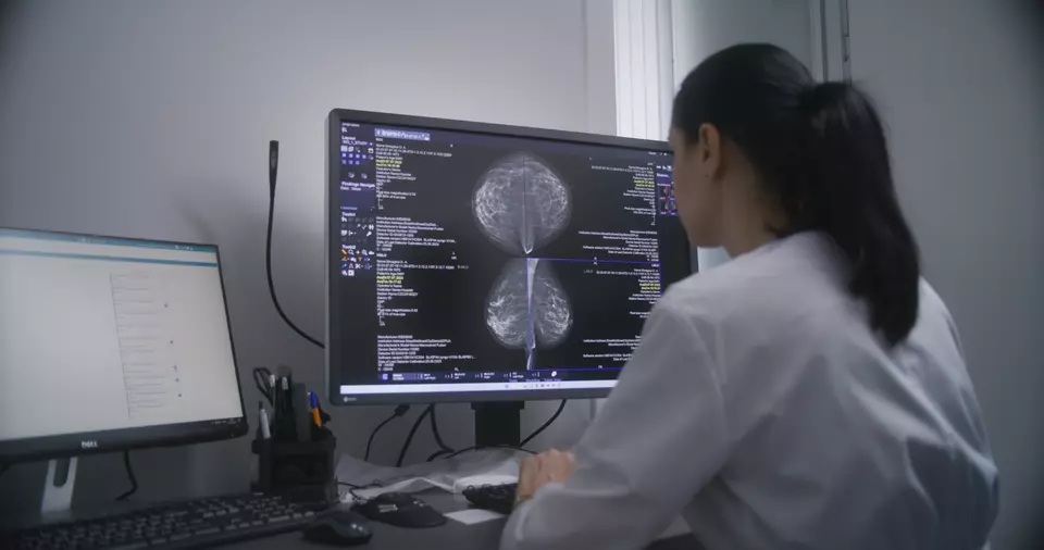 A female doctor wearing a white coat faces a computer. On the computer screen is a mammogram image. 