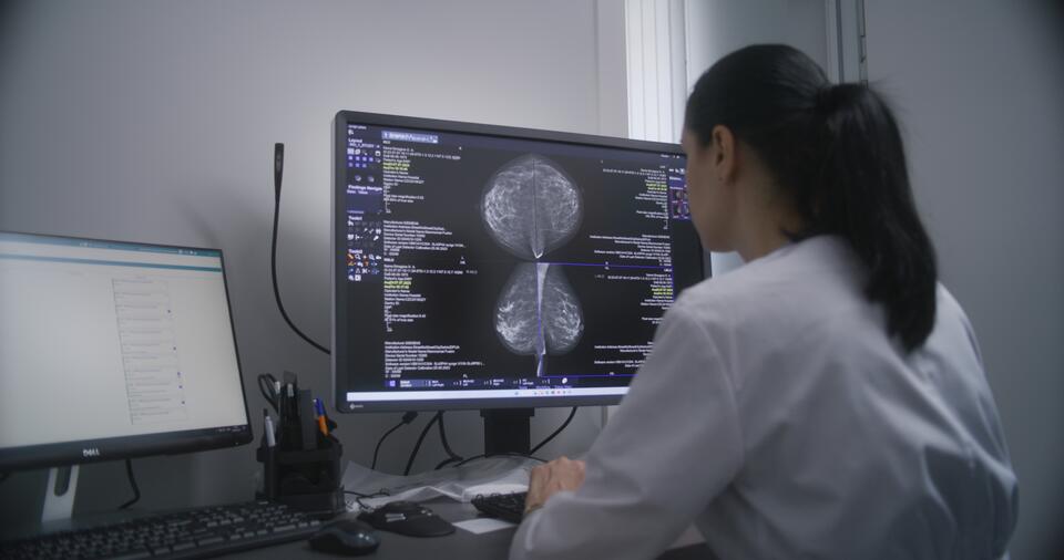 A female doctor wearing a white coat faces a computer. On the computer screen is a mammogram image. 