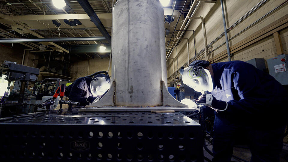 Two people wearing welding masks and other safety equipment bend over a table to work on a large metal cylinder.