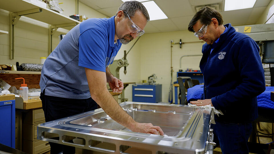 Two researchers wearing safety glasses lean over a rectangular metal case lying flat on a table.
