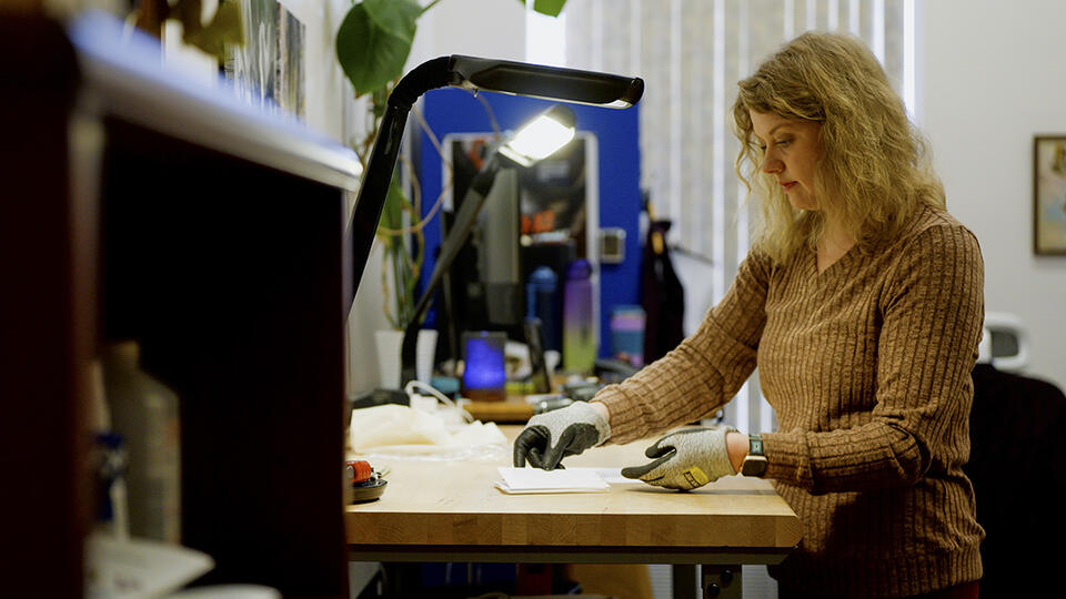 A woman wearing special gloves stands at a high table, sorting small pieces of paper. 