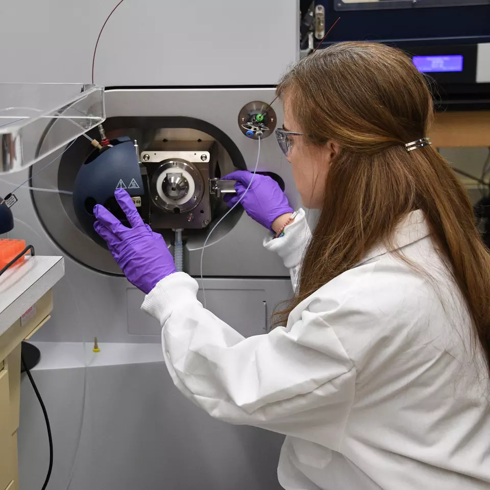 A woman wearing purple gloves and a lab coat opens the cover of a large piece of scientific equipment.