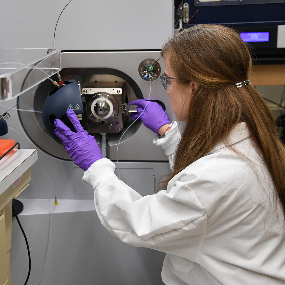 A woman wearing purple gloves and a lab coat opens the cover of a large piece of scientific equipment.