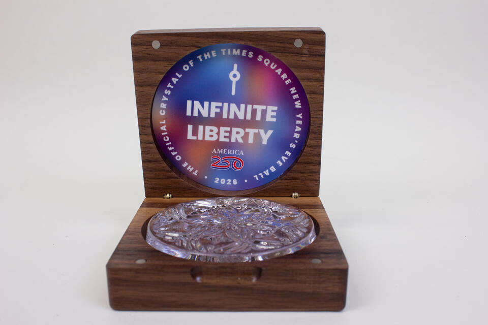 A clear crystal sits in a wooden box. On the top center of the box is a label that reads  "The Official Crystal of The Times Square New Year's Eve Ball 2026"