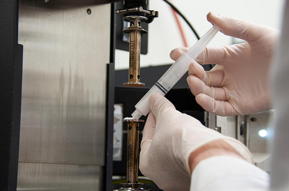 Researcher holds a small white tube filled with wet ceramics and points it toward lab equipment.