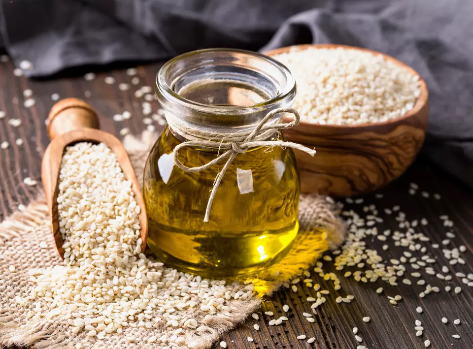 Bottle of sesame oil on a wooden table.  Sesame seeds are in a bowl and spoon next to the bottle and also sprinkled on the table.